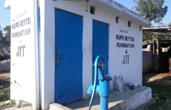 A bathroom with two blue doors. A blue water pump sits in front of the bathroom. There is a sign on the bathroom that says, "Supported by the Hope Better Foundation.