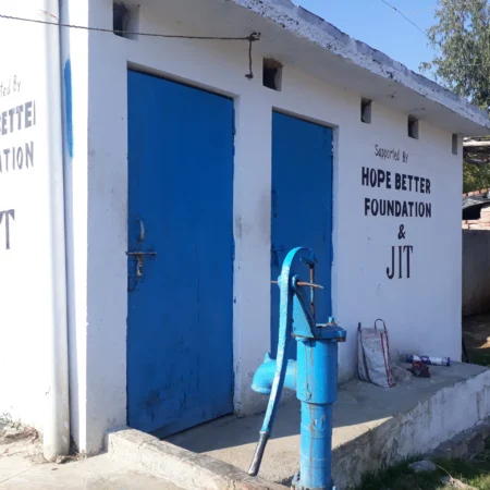 A bathroom with two blue doors. A blue water pump sits in front of the bathroom. There is a sign on the bathroom that says, "Supported by the Hope Better Foundation.