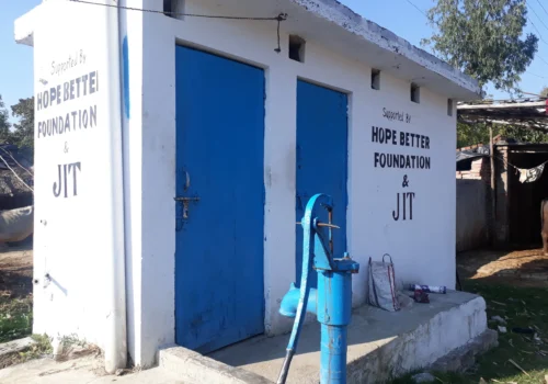 A bathroom with two blue doors. A blue water pump sits in front of the bathroom. There is a sign on the bathroom that says, "Supported by the Hope Better Foundation.