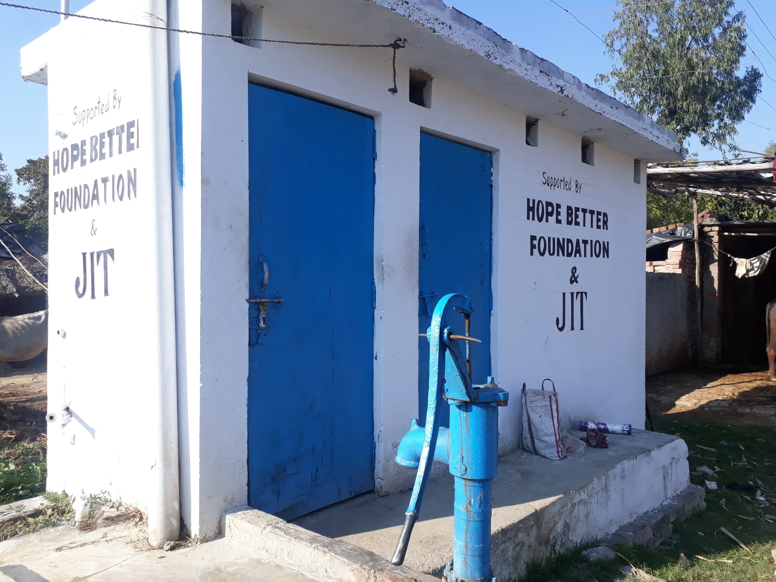 A bathroom with two blue doors. A blue water pump sits in front of the bathroom. There is a sign on the bathroom that says, "Supported by the Hope Better Foundation.